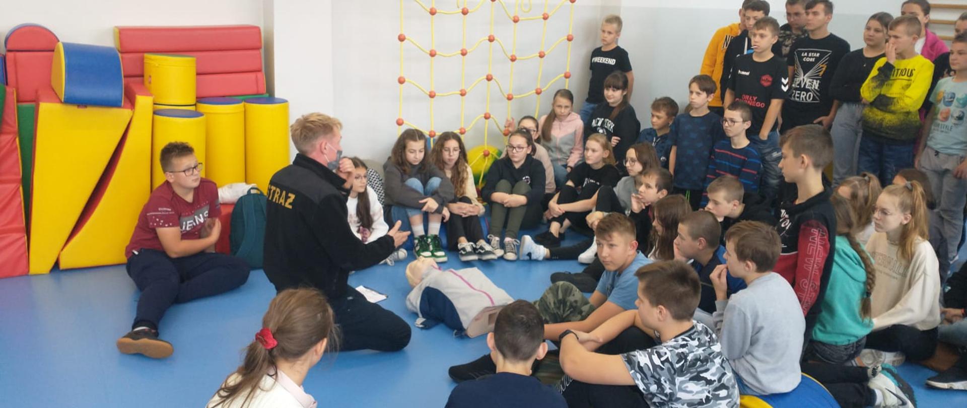 Strażak klęczy przed manekinem Naokoło dzieci przysłuchują się . Zdjęcie na sali gimnastycznej 