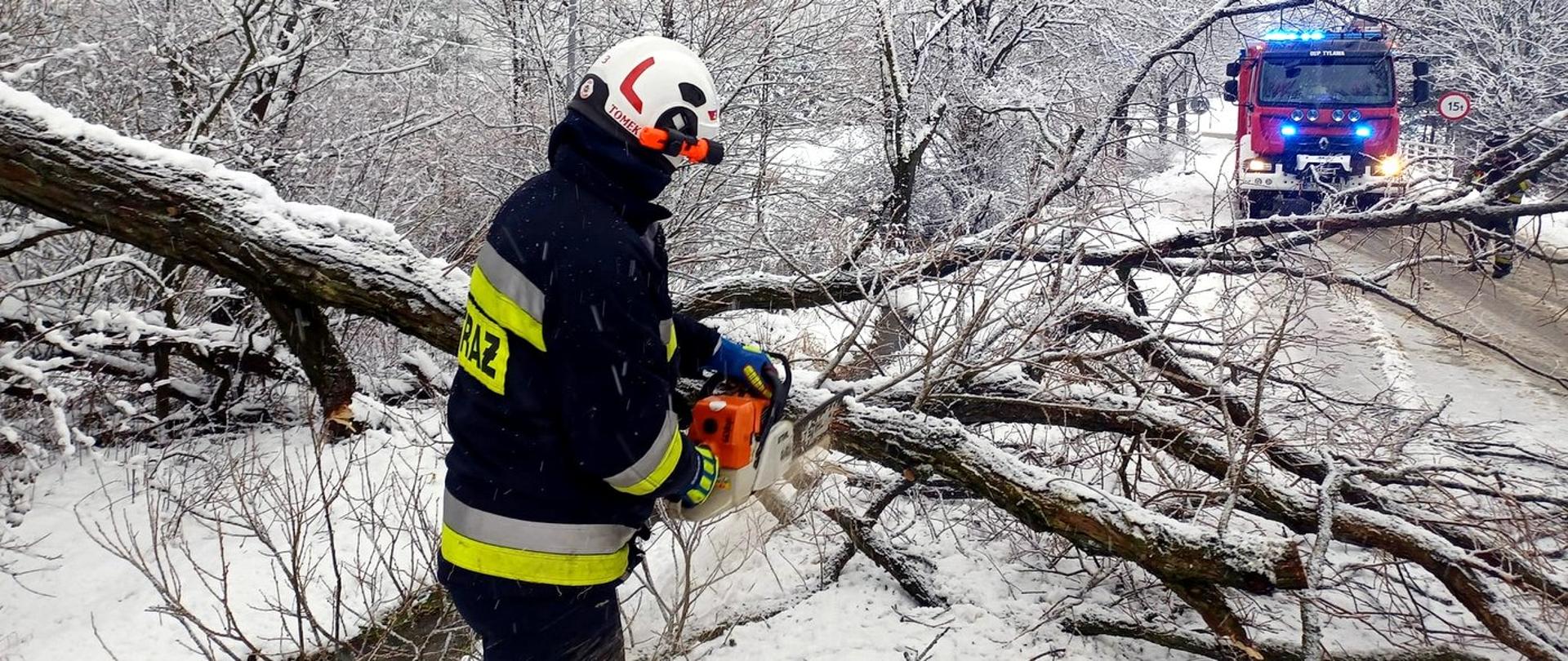 Zdjęcie zrobione na zewnątrz. Strażak, używając pilarki spalinowej, usuwa drzewo, które runęło na jezdnię. W tle stoi samochód pożarniczy.