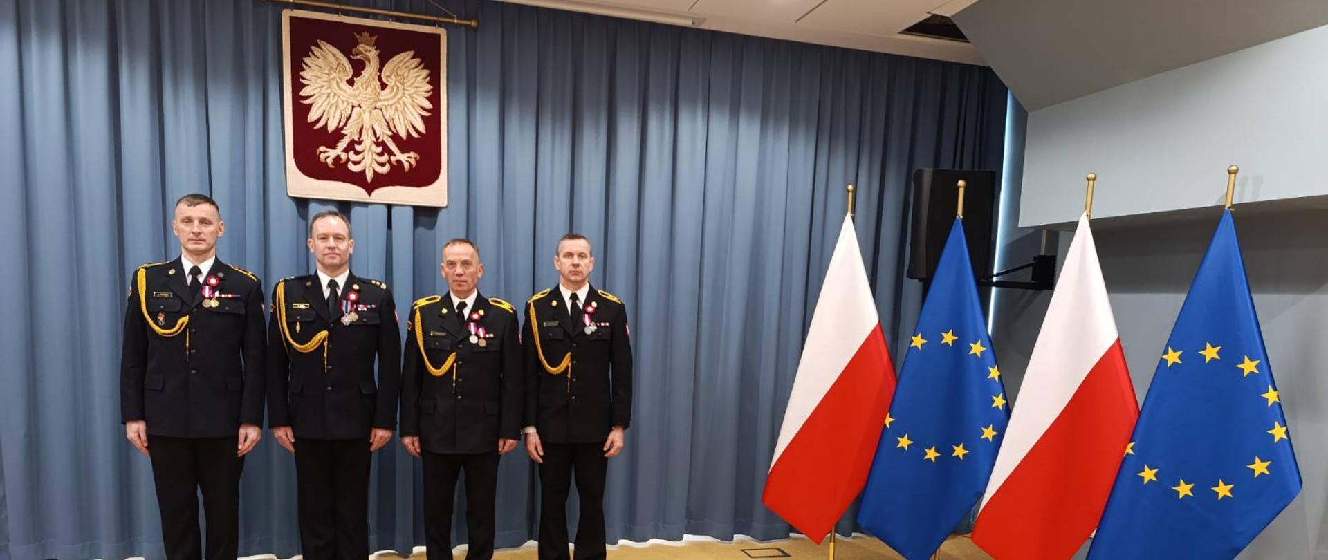 Zdjęcie przedstawia 4 funkcjonariuszy z KP PSP Sokołów Podlaski w budynku KW PSP Warszawa. Ubrani w mundury galowe koloru czarnego z odznaczeniami ( medalami ) na piersi. W tle godło Polski, po prawek stronie na przemian 2 flagi Polski i Unii Europejskiej.
