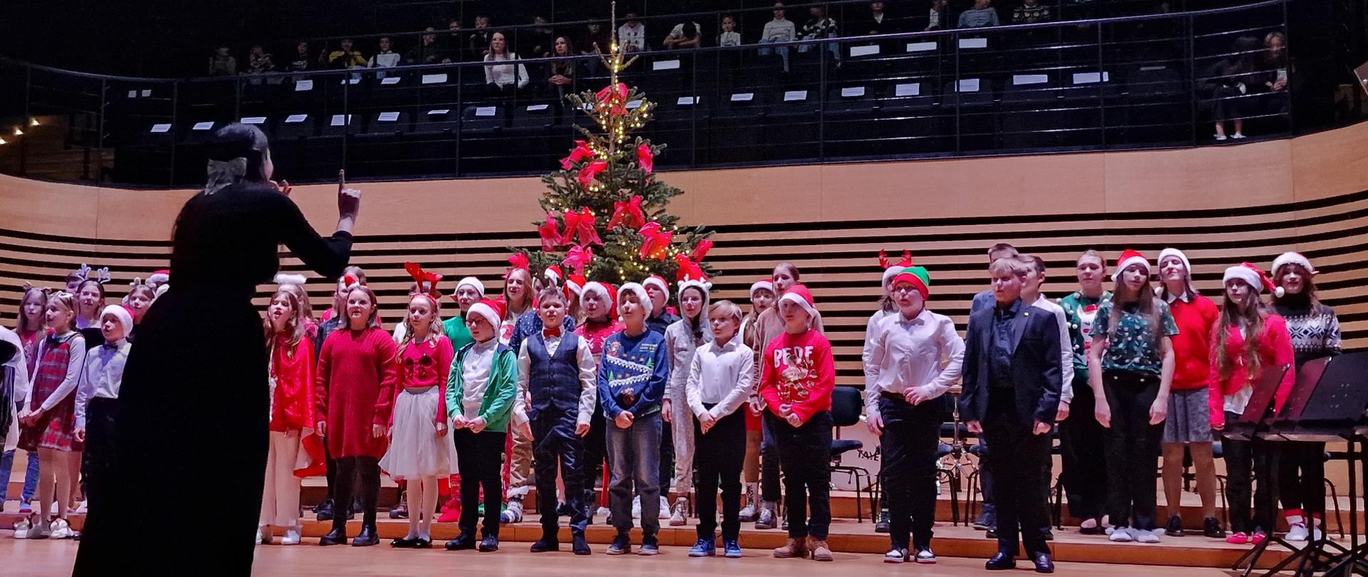 Grupa ok.35 dzieci, ubranych w kolorowe świąteczne stroje stoi w trzech rzędach na estradzie sali koncertowej i śpiewa. Przed nimi, tyłem do widowni stoi kobieta i dyryguje.