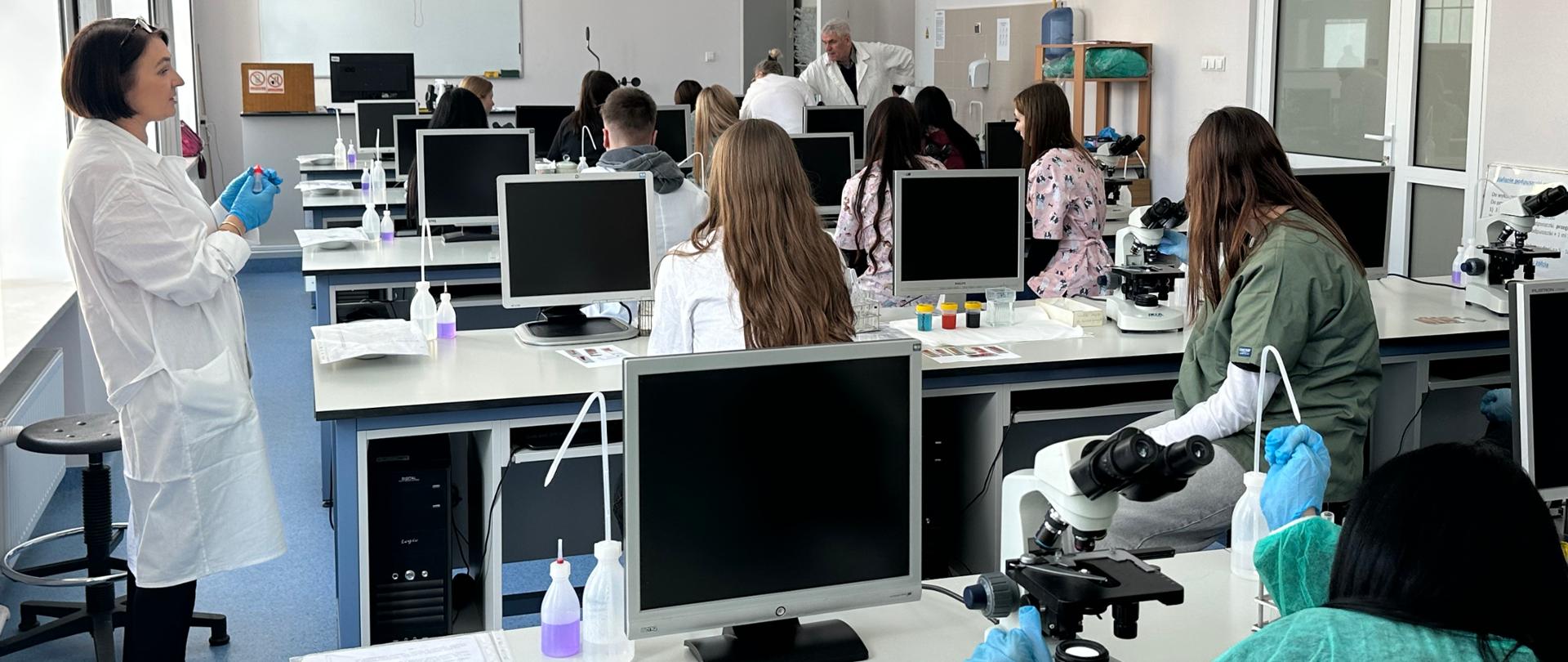 Uczniowie siedzą przy stanowiskach laboratoryjnych słuchając wykładu osoby prowadzącej