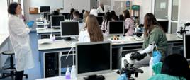 Uczniowie siedzą przy stanowiskach laboratoryjnych słuchając wykładu osoby prowadzącej