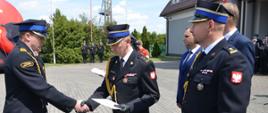 Na zdjęciu widać pięć osób. Trzy osoby ubrane są w mundury galowe ze złotym sznurem a dwóch mężczyzna w niebieskie garnitury. Jeden ze strażaków trzyma w ręku białą teczkę, którą wręcza drugiemu strażakowi. W tle widać maszt biało-czerwony, przed którym stoją trzej strażacy w mundurach galowych ze złotym sznurem. Na głowach mają czapki. Widać również dmuchany baner w kształcie hełmu strażackiego, częściowo budynek komendy, przed którym stoją strażacy z ochotniczej straży pożarnej oraz krzewy. Jest widno.