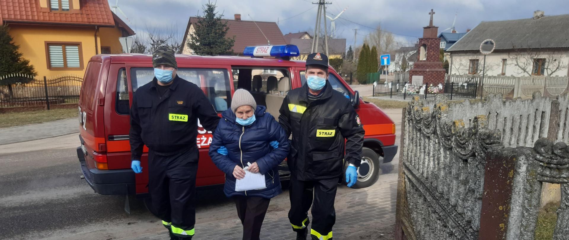 Dwóch strażaków OSP prowadzi starszą kobietą z samochodu osobowego strażackiego do domu.