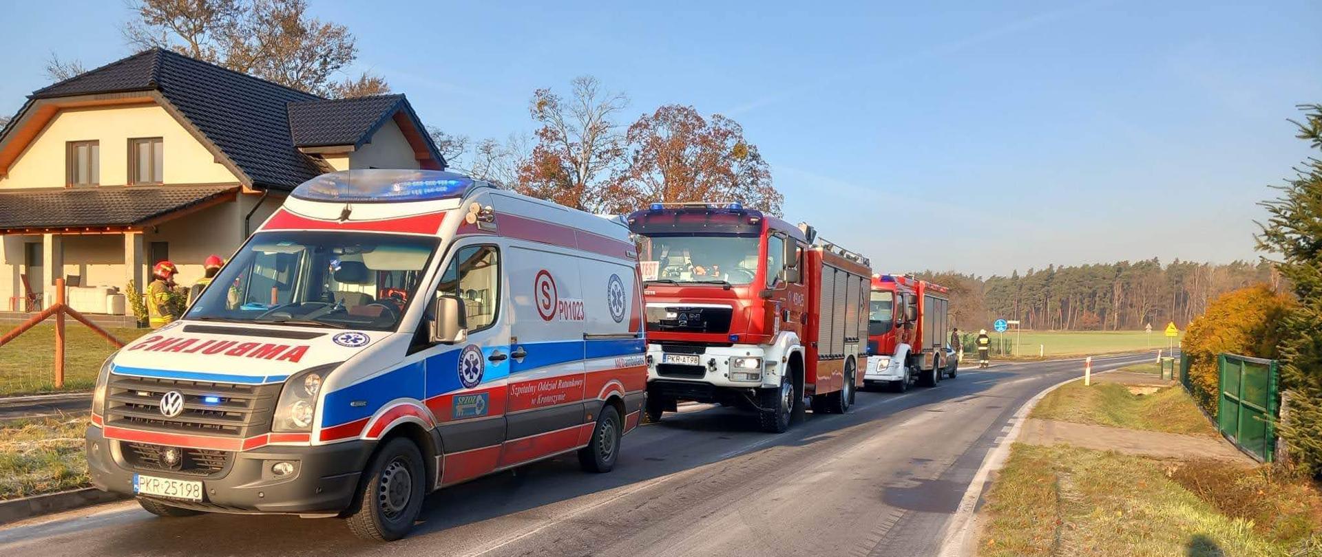 Na drodze stoi karetka pogotowia oraz dwa pojazdy straży pożarnej. Trwają działania związane z wypadkiem komunikacyjnym. W oddali widać strażaków w ubraniach bojowych realizujących czynności. W tle zabudowania oraz obręb leśny