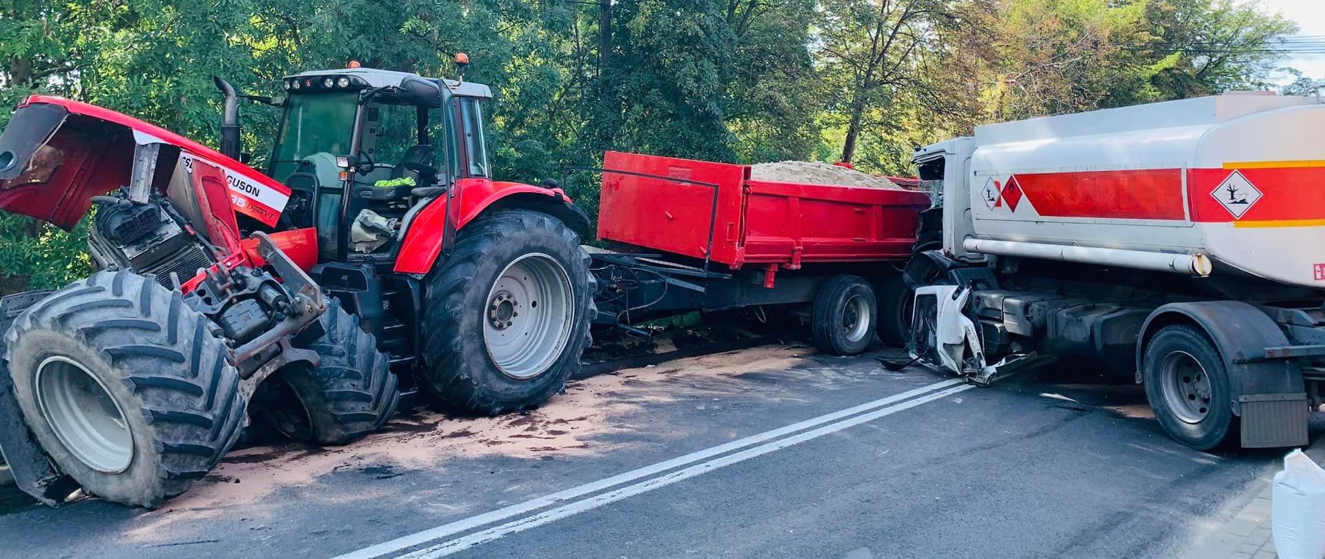 Po lewej stronie rozbity ciągnik rolniczy z przyczepą wypełnioną piaskiem. Pod nim przy krawężniku plama ropopochodna. Po prawej stronie samochód ciężarowy do przewozu paliw po uderzeniu w przyczepę ciągniętą przez ciągnik rolniczy.