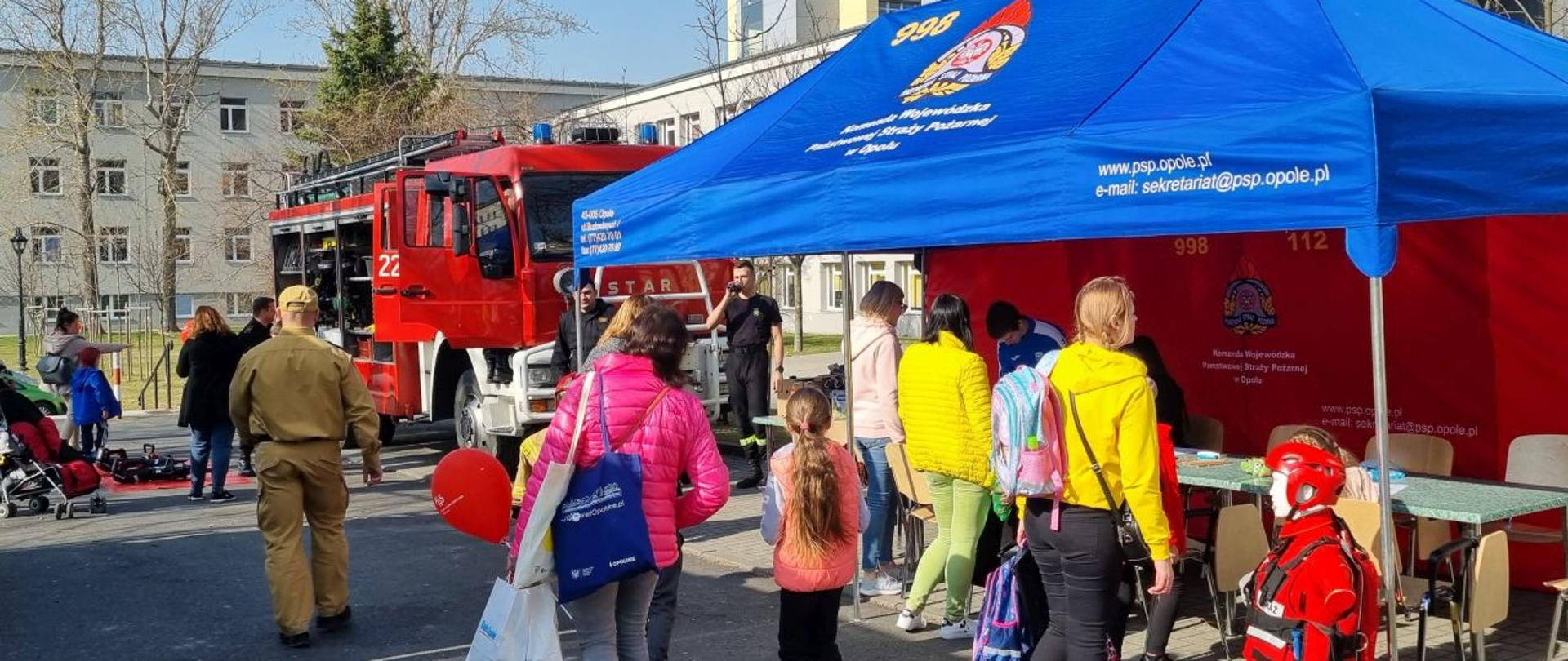 Dzieci, młodzież i dorośli stojące przy i pod namiotem Komendy Wojewódzkiej PSP w Opolu oraz dzieci i dorośli stojący przy samochodzie ratowniczo-gaśniczym (w oddali) podczas oglądania sprzętu i samego samochodu. W tle widoczny kompleks Uniwersytetu Opolskiego.