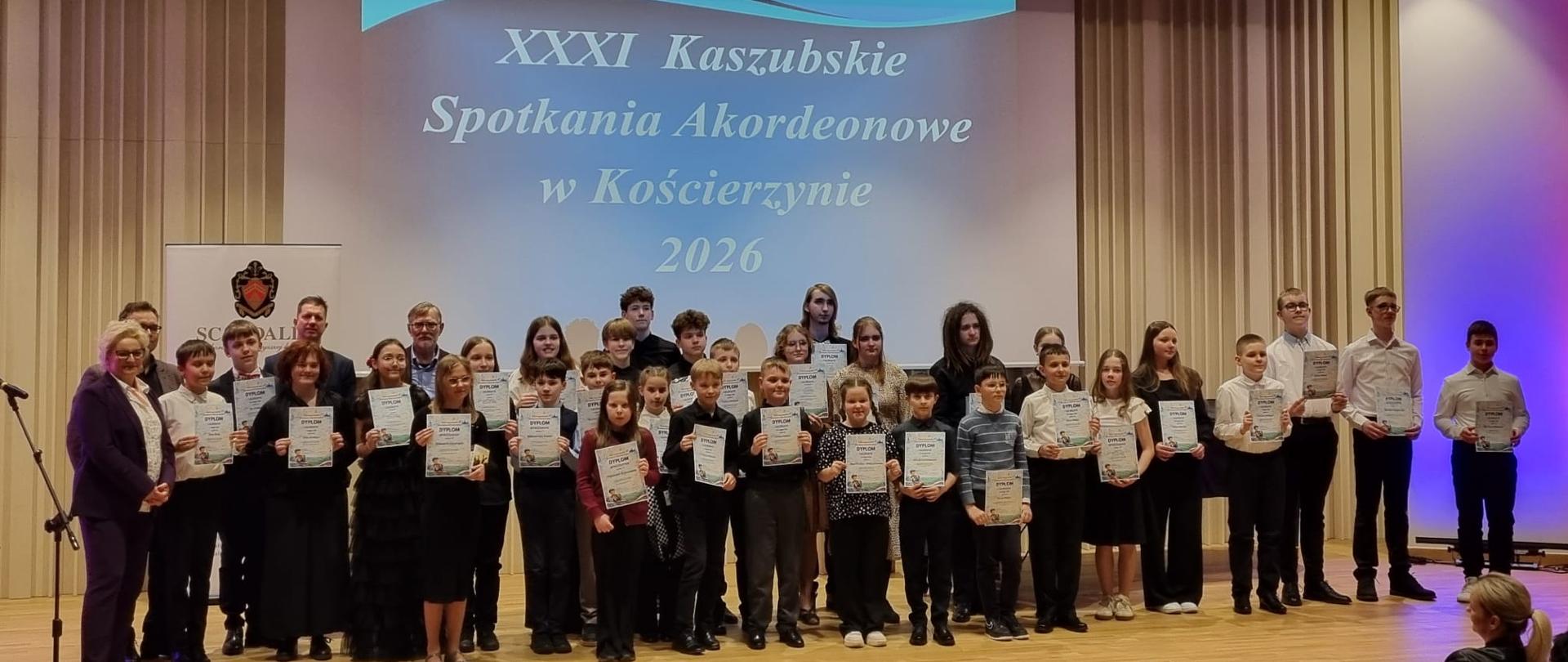 Na zdjęciu odświętnie ubrani uczniowie, którzy zostali laureatami XXXI Kaszubskich Spotkań Akordeonowych. Zdjęcie zostało wykonane podczas wręczenia nagród. Tło obrazka w odcieniach beżu. W tle na ścianie rzut niebieskiego slajdu z białymi napisem "XXXI Kaszubskie Spotkania Akordeonowe w Kościerzynie 2026". W oddali czarny fortepian, przed sceną zgromadzona publiczność.