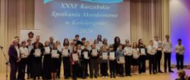 Na zdjęciu odświętnie ubrani uczniowie, którzy zostali laureatami XXXI Kaszubskich Spotkań Akordeonowych. Zdjęcie zostało wykonane podczas wręczenia nagród. Tło obrazka w odcieniach beżu. W tle na ścianie rzut niebieskiego slajdu z białymi napisem "XXXI Kaszubskie Spotkania Akordeonowe w Kościerzynie 2026". W oddali czarny fortepian, przed sceną zgromadzona publiczność.