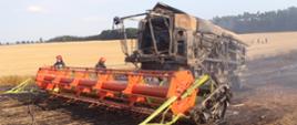 Zdjęcie przedstawia spalony kombajn zbożowy marki Class, znajdujący się na spalonym ściernisku. W tle znajduje się las, bezchmurne niebo oraz w oddali strażacy. Zdjęcie wykonano za dnia
