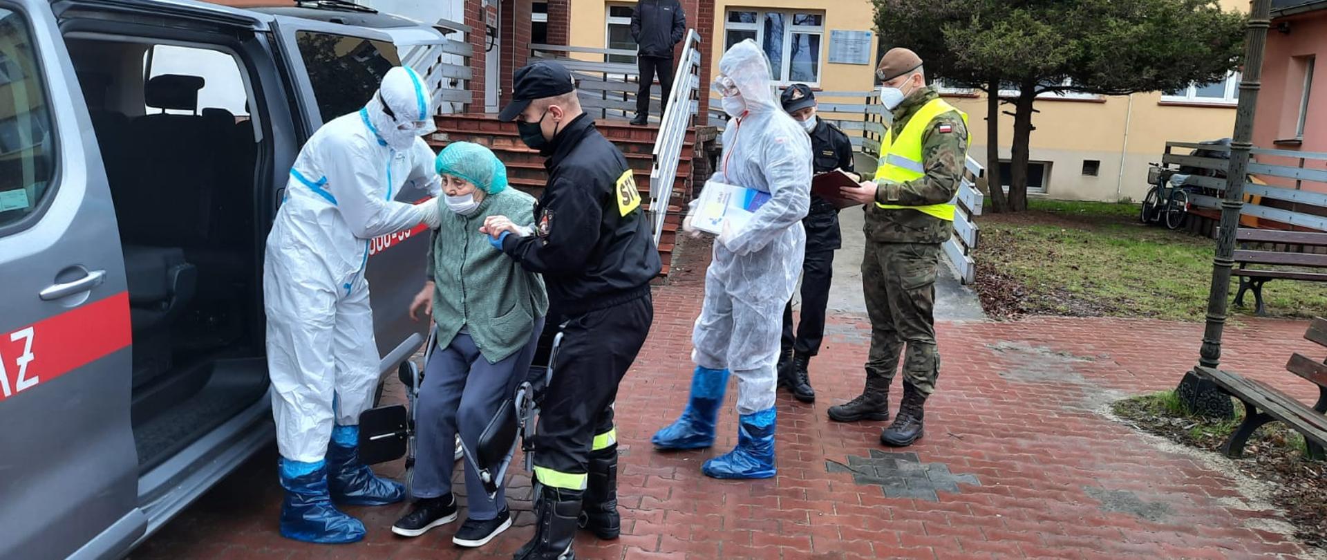 Fotografia przedstawia ujęcie przed budynkiem szpitala. Strażak w ubraniu koszarowym z założoną maseczką i rękawiczkami jednorazowymi, wraz z pracownikiem szpitala ubranym w kombinezon, gogle, rękawiczki jednorazowe oraz ochraniacze obuwia pomagają starszej kobiecie wstać z wózka inwalidzkiego i wsiąść do busa strażackiego. Dalej widoczny jeszcze jeden pracownik szpitala ubrany jak ten pierwszy, za nim oficer PSP w ubraniu dowódczo-sztabowym konsultuje coś z żołnierzem Wojsk Obrony Terytorialnej, który także ma założoną maseczkę oraz trzyma w ręku notatki. Ujęcie z lewej strony busa, jest dzień, pochmurno.