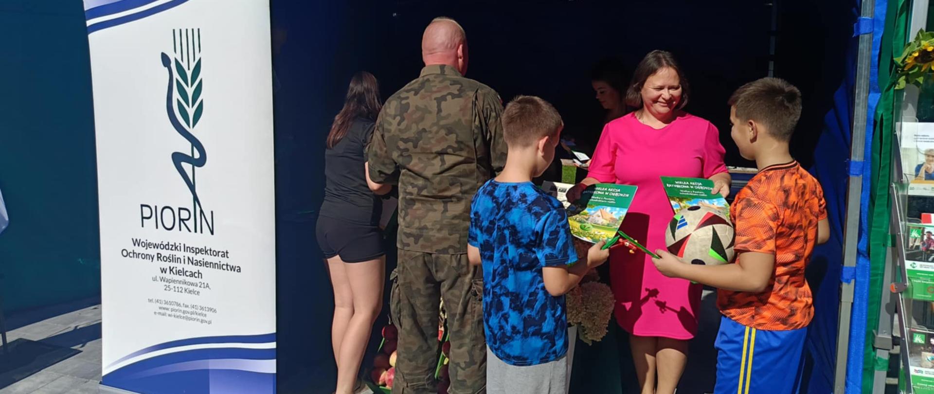 Kobieta pracownik Wojewódzkiego Inspektoratu Ochrony Roślin i Nasiennictwa w Kielcach Oddział w Sandomierzu rozdaje zgromadzonym dzieciom gadżety promujące Inspekcje oraz kolorowanki. 