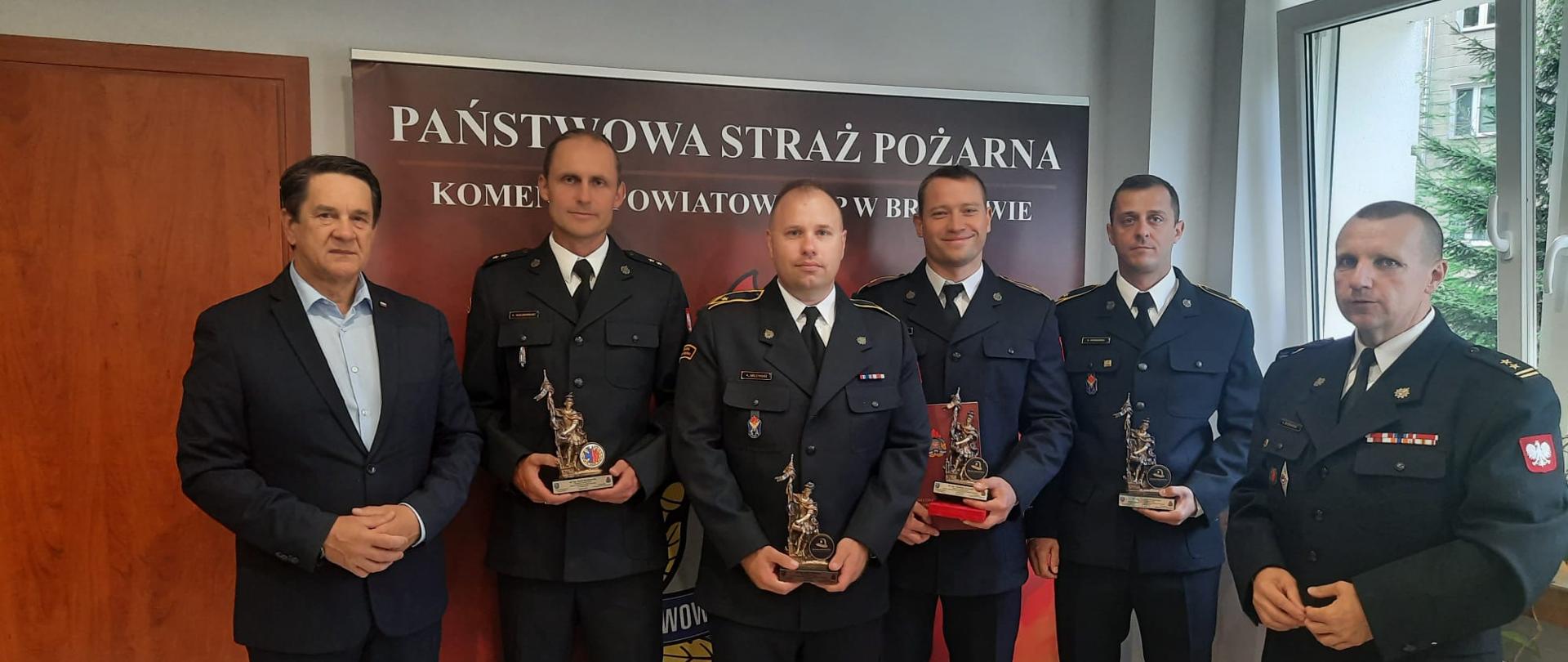 Na zdjęciu na tle ścianki z napisem Państwowa Straż Pożarna stoi czterech wyróżnionych strażaków w mundurach galowych ze statuetkami w ręku. Dodatkowo z prawej strony stoi Komendant powiatowy a z lewej Starosta braniewski.