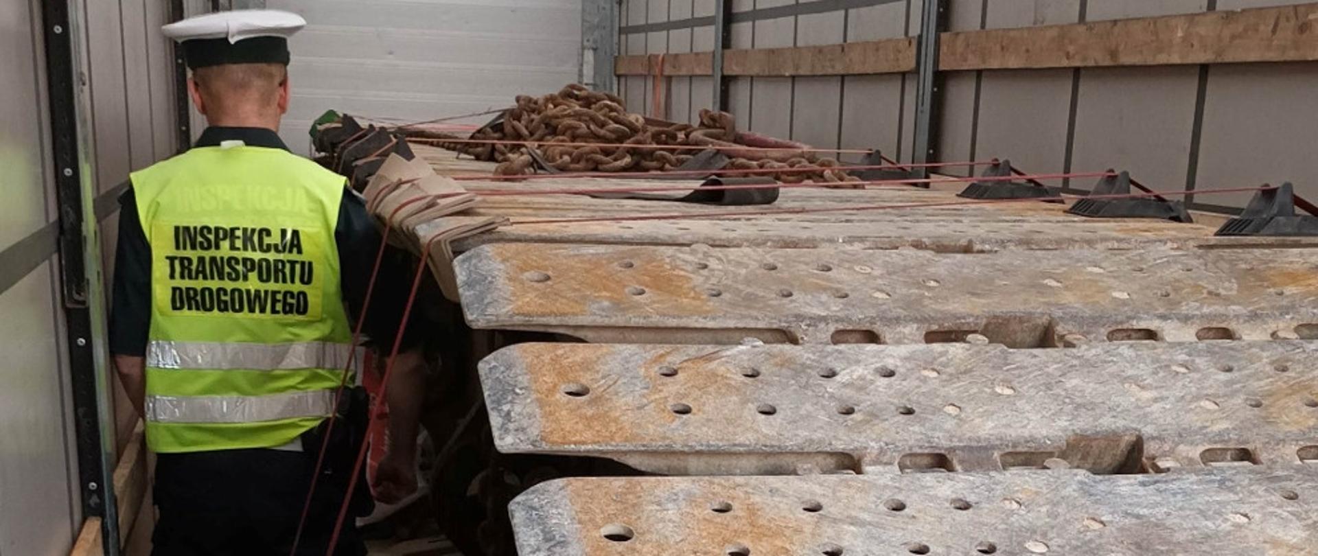 Inspektor ITD stoi w przestrzeni ładunkowej naczepy ciężarowej i kontroluje prawidłowość zabezpieczenia przewożonej gąsienicy do maszyny budowlanej.