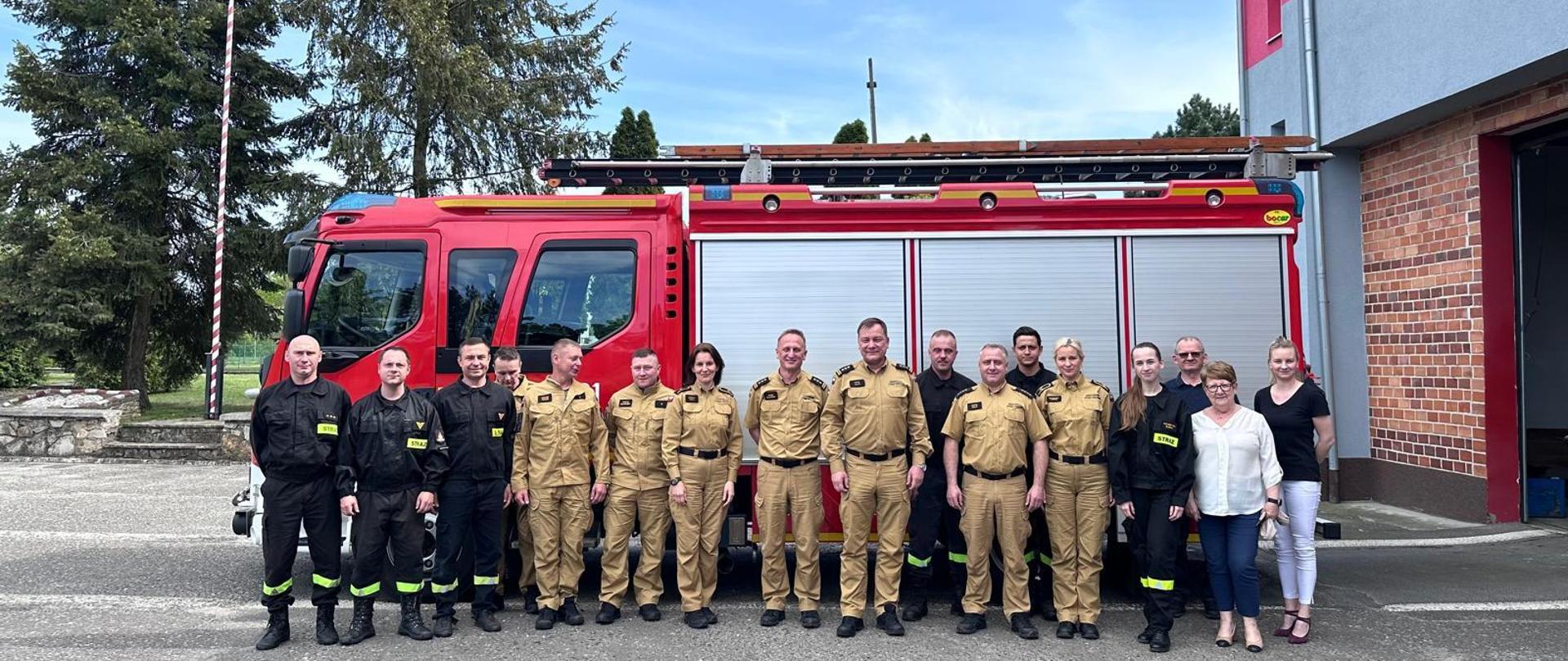 Wspólne zdjęcie strażaków i pracowników cywilnych Komendy Powiatowej Państwowej Straży Pożarnej w Strzelcach Opolskich z Opolskim Komendantem Wojewódzkim Państwowej Straży Pożarnej st. bryg. Pawłem Kielarem. Załoga strzeleckiej komendy wraz z komendantem stoją na placu przed komendą na tle czerwonego pojazdu pożarniczego. W tle widoczna Flaga Rzeczypospolitej Polskiej na maszcie.