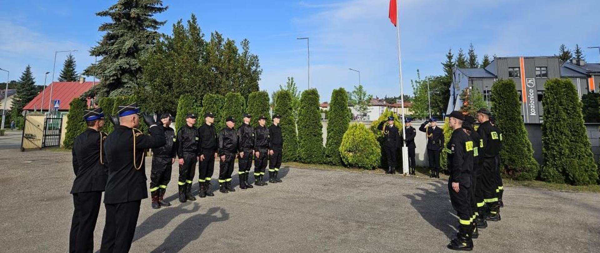 strażacy ze zmian służbowych z dowódcą JRG i komendantem stoją na pacu przy maszcie flagowym podczas uroczystego podniesienia flagi