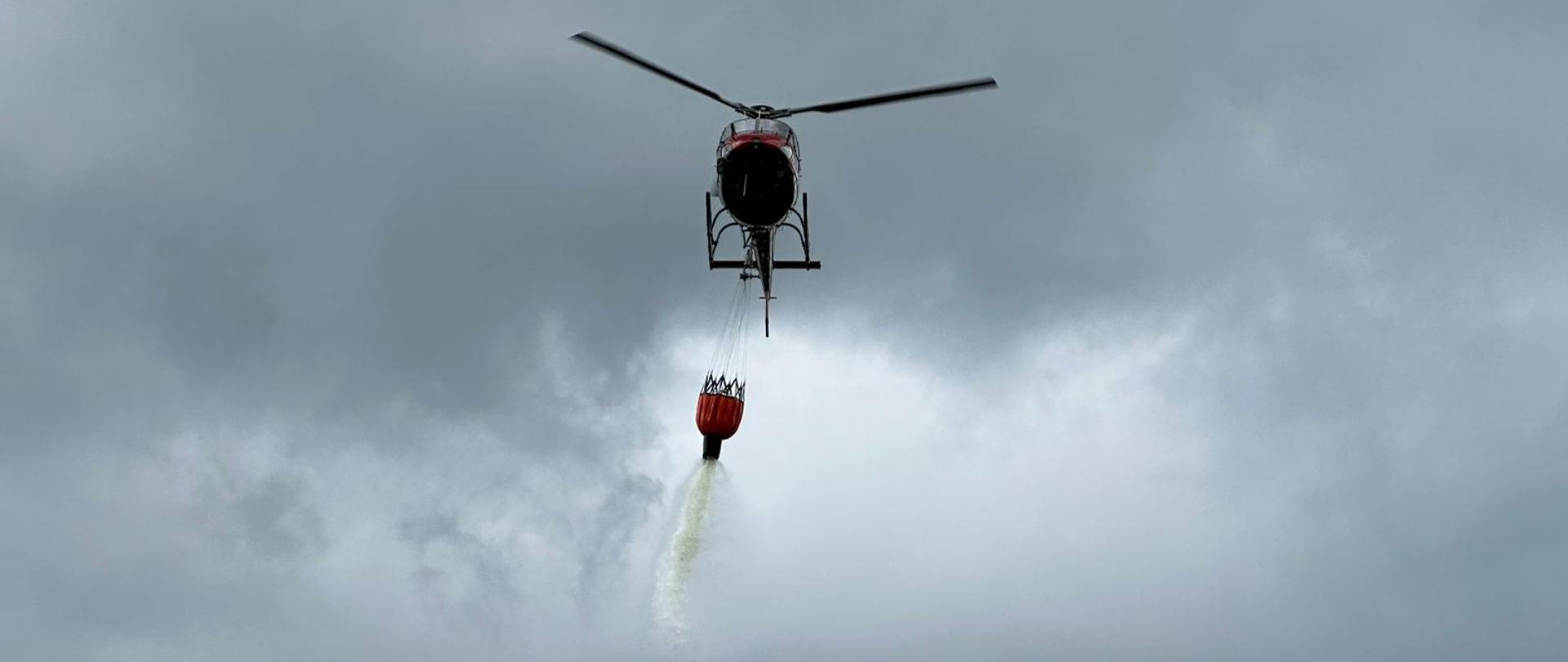 Śmigłowiec Lasów Państwowych dokonuje zrzutu. Woda wydostaje się z podwieszonego pod statek powietrzny specjalnego zbiornika typu bambi bucket.