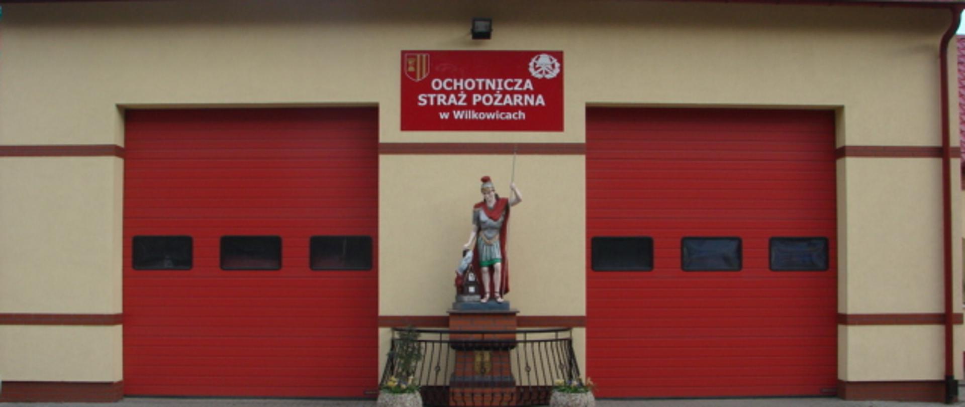 Widoczny budynek z dwiema zamkniętymi bramami garażowymi oraz figurą św. Floriana i czerwoną tablicą z białym napisem.