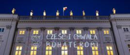 Pałac Prezydencki w Warszawie. Zdjęcie wykonane w nocy. W zabytkowym, klasycystycznym budynku świecą się światła w nocy. Na środku zdjęcia grafika - szaro-biały orzełek Polski i szaro-białe napisy: "Cześć i chwała bohaterom".