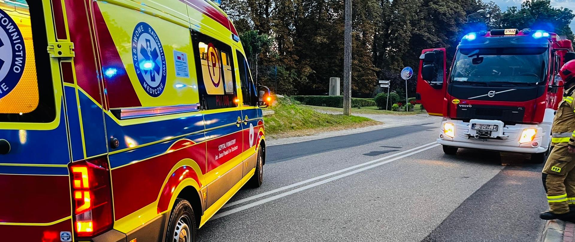 Motocykl przewrócony na jezdni po wypadku. Obok stoi karetka pogotowia i wóz straży pożarnej z włączonymi światłami ostrzegawczymi.