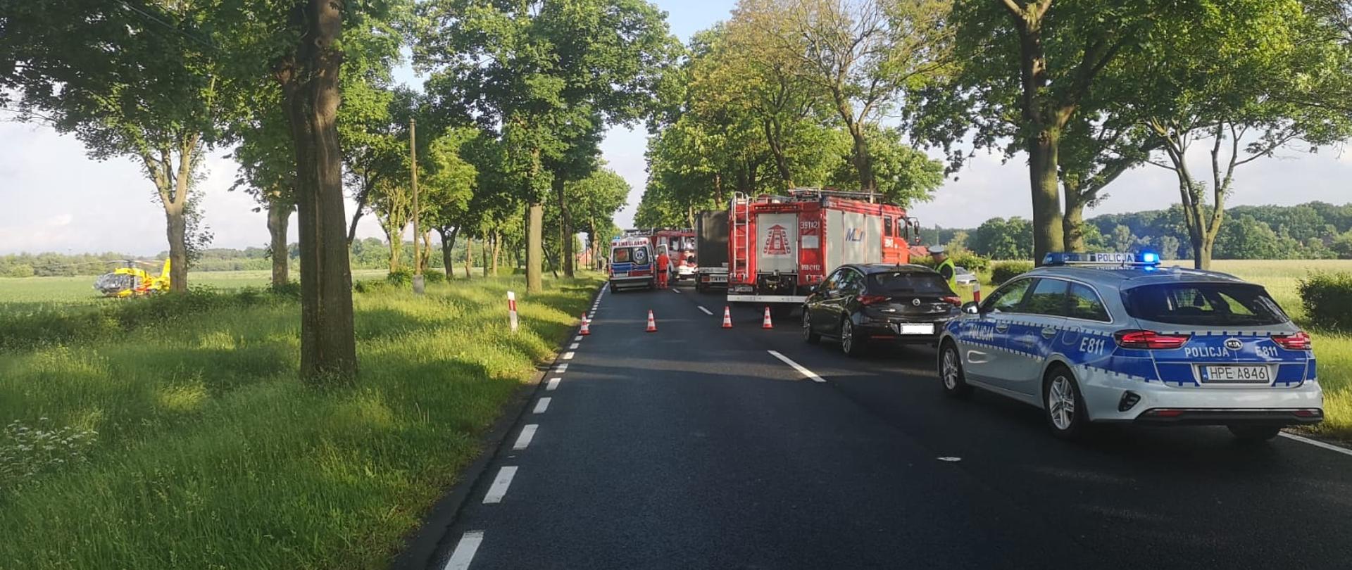 Widok stojących na jezdni pojazdów służb tj. straży pożarnej, policji oraz pogotowia ratunkowego. Z lewej strony na trawie stoi śmigłowiec LPR.