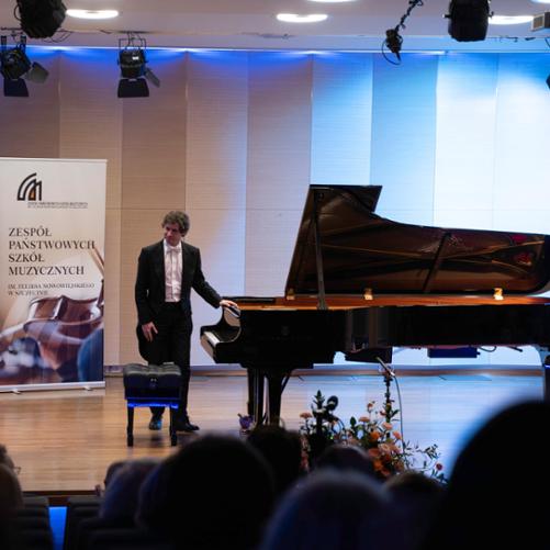 zdjęcie przedstawia pianistę kłaniającego się przy fortepianie na scenie sali koncertowej przed rozpoczęciem koncertu, w tle banery szkoły i sponsorów