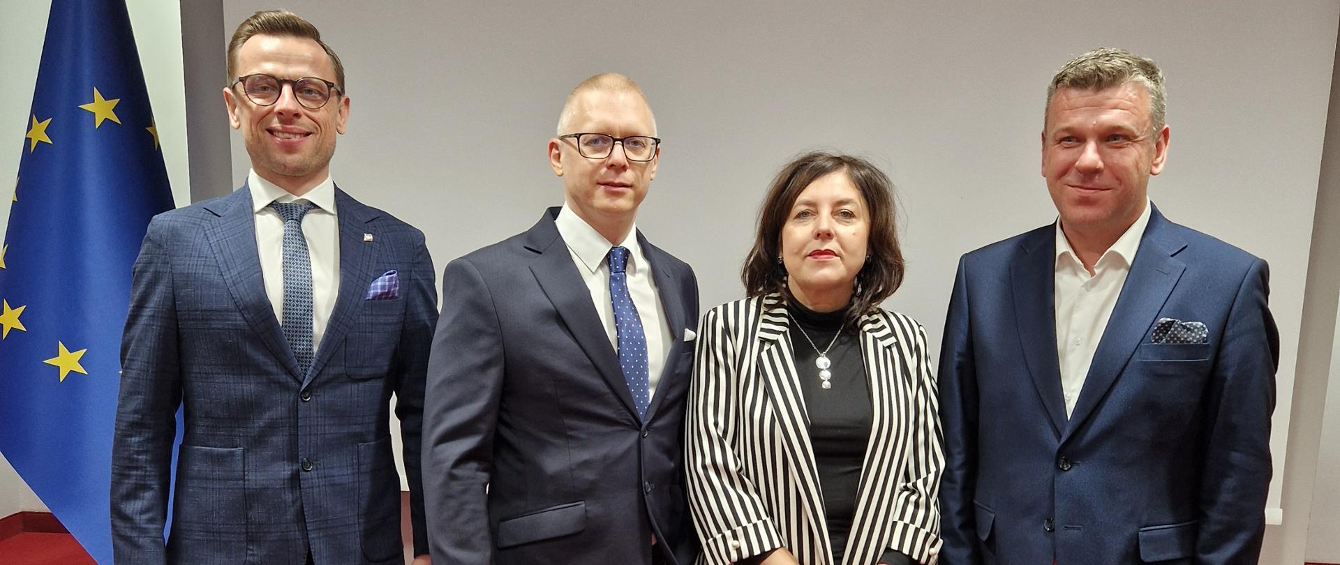 Grupa czterech osób pozuje do formalnego zdjęcia na tle flagi Unii Europejskiej. Dwóch mężczyzn stojących po lewej stronie ma na sobie ciemne garnitury i niebieskie krawaty, obaj w jasnych koszulach. Między nimi stoi kobieta w czarno-białym, pasiastym stroju, a obok niej znajduje się mężczyzna w niebieskim garniturze.