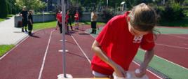 Widok z przodu. Zawodniczka w czerwonej koszulce w trakcie wyciągania z pudełka piłeczki do tenisa stołowego w ramach konkurencji „ping-pongowy slalom”. W tle pozostali członkowie drużyny w oczekiwaniu na swój bieg. 