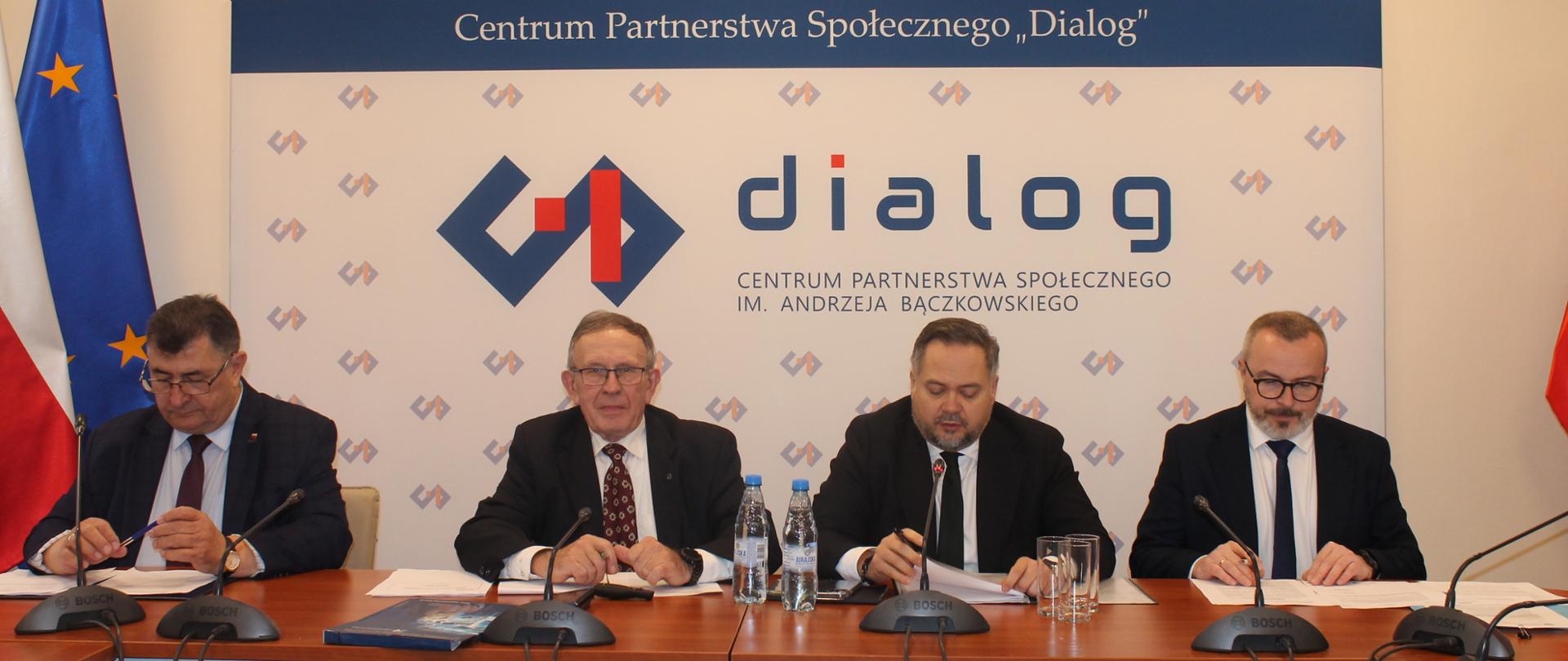 Na zdjęciu widnieje stół prezydialny za którym siedzą: Pan Roman Jakim, Współprzewodniczący Zespołu ze strony związków zawodowych; Pan Marek Dras, Współprzewodniczący Zespołu ze strony pracodawców; Pan Konrad Gołota, Podsekretarz Stanu w Ministerstwie Aktywów Państwowych oraz Pan Marcin Kubicza, Dyrektor Departamentu Projektów Strategicznych w Ministerstwie Aktywów Państwowych. W tle znajduje się baner z napisem Centrum Partnerstwa Społecznego „Dialog” im. Andrzeja Bączkowskiego. Po lewej stronie banneru znajdują się dwie flagi – Polski oraz Unii Europejskiej. Nad banerem znajduje się Godło Polski.