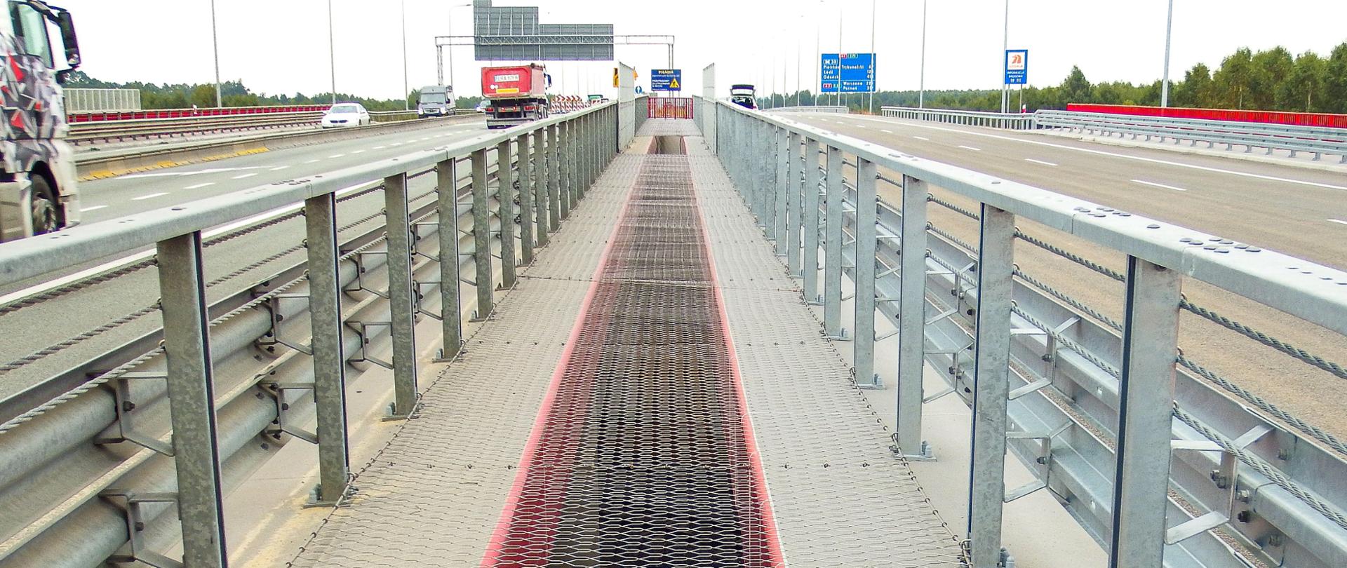 Fotografia z poziomu terenu na zabezpieczone siatką odstępy pomiędzy pomostami wiaduktów autostrady A1. Wiadukty zlokalizowane są na obu jezdniach autostrady, po których odbywa się ruch pojazdów. Belki gzymsowe obiektów w kolorze czerwonym. 
