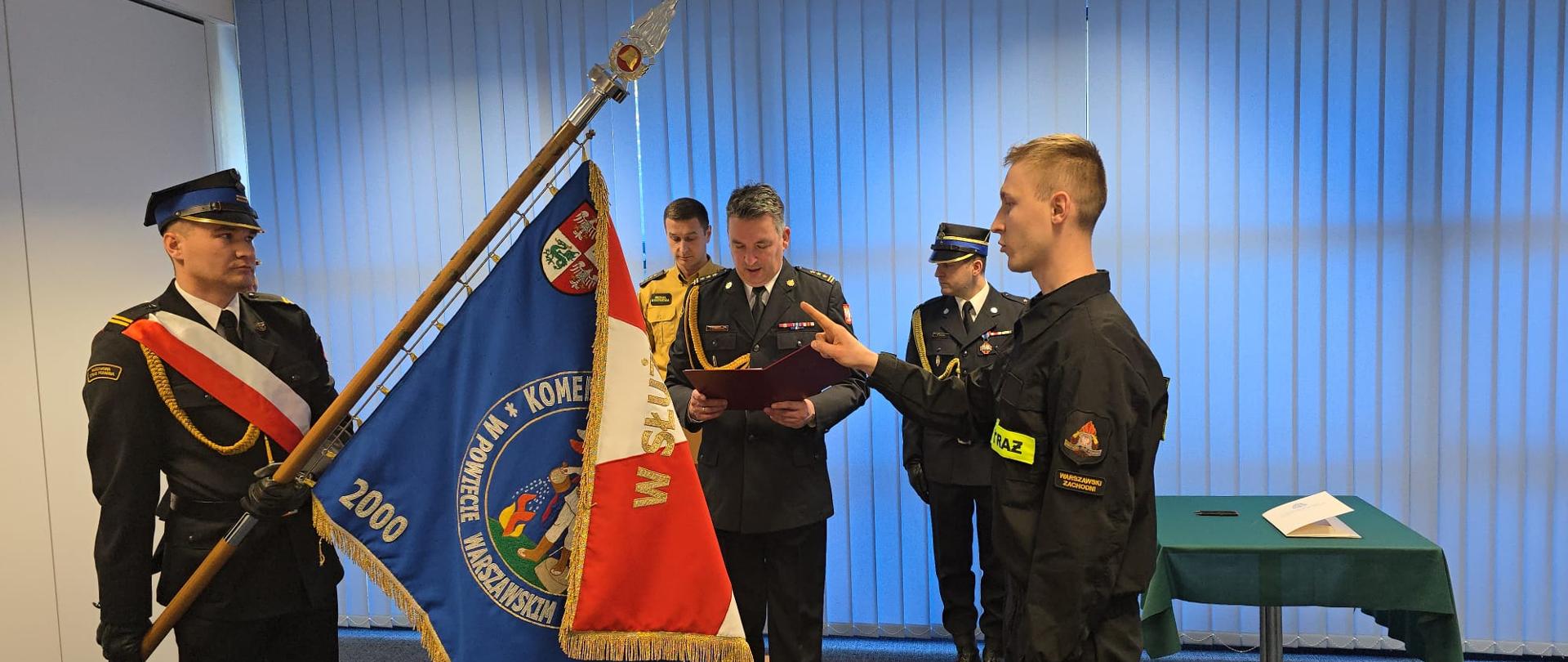 Zdjęcie przedstawia oficjalną uroczystość o charakterze państwowym lub służbowym, prawdopodobnie związaną ze strażą pożarną w Polsce.
Oto szczegółowy opis tego, co znajduje się na obrazku:
Postacie i ich role
• Główny bohater (po prawej): Młody mężczyzna w czarnym mundurze ćwiczebnym z napisem "STRAŻ" na piersi. Stoi on w postawie wyprostowanej z podniesioną dłonią w geście składania przysięgi (dwa palce wyciągnięte ku górze).
• Osoba prowadząca (w centrum): Oficer w ciemnym mundurze wyjściowym z dystynkcjami i baretkami (odznaczeniami) na piersi. Odczytuje on treść przysięgi lub rotę z czerwonej teczki.
• Poczet sztandarowy (po lewej): Strażak w pełnym umundurowaniu wyjściowym z biało-czerwoną szarfą, trzymający pochylony sztandar.
• Tło: W tle widać dwóch innych funkcjonariuszy w mundurach, którzy obserwują ceremonię.
Symbolika i detale
• Sztandar: Jest niebieski z bogatymi haftami. Widnieje na nim napis związany z Komendą Powiatową w Powiecie Warszawskim Zachodnim oraz daty „2000” i „2010”. Na drzewcu zamontowany jest ozdobny grot.
• Otoczenie: Uroczystość odbywa się w pomieszczeniu biurowym lub sali konferencyjnej. Za plecami uczestników znajdują się jasnoniebieskie żaluzje pionowe (verticale). Po prawej stronie stoi stolik przykryty zielonym obrusem, na którym leżą dokumenty.
Atmosfera
Zdjęcie emanuje powagą i dyscypliną. Jest to klasyczny moment ślubowania nowego funkcjonariusza, co podkreśla obecność sztandaru i galowych mundurów.
