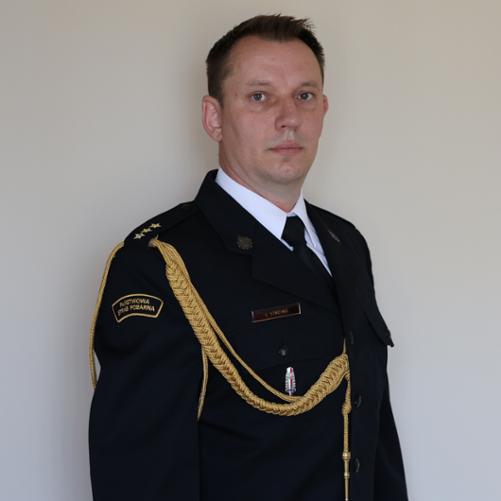 Zdjęcie kapitana Łukasza Staciwy w mundurze galowym Państwowej Straży Pożarnej. Stoi na tle jasnej ściany, zwrócony lekko bokiem, patrząc w obiektyw. Mundur zdobią złote sznury reprezentacyjne oraz odznaczenia służbowe