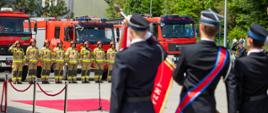 Na zdjęciu widoczni strażacy zmiany służbowej w umundurowaniu piaskowym specjalnym w czerwonych hełmach. Od tyłu widoczni strażacy OSP ze sztandarem w umundurowaniu galowym granatowym w czapkach rogatywkach podczas Powiatowych obchodów Dnia Strażaka w KP PSP w Gorlicach.