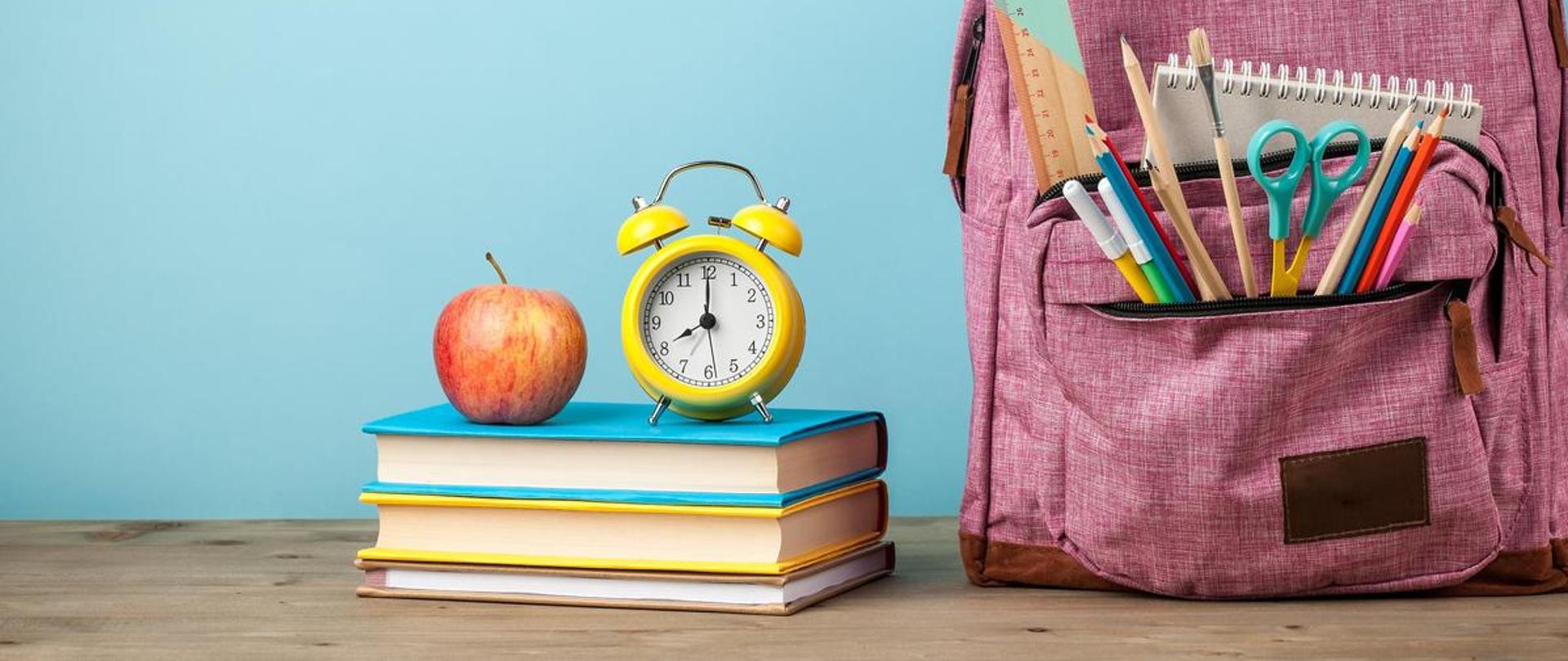 Na niebieskim tle po prawej stronie widzimy różowy plecak szkolny wypełniony przyborami szkolnymi oraz zeszytami. W centralnej części widzimy 3 podręczniki szkolne na których ustawiony jest zegarek, obok którego leży jabłko