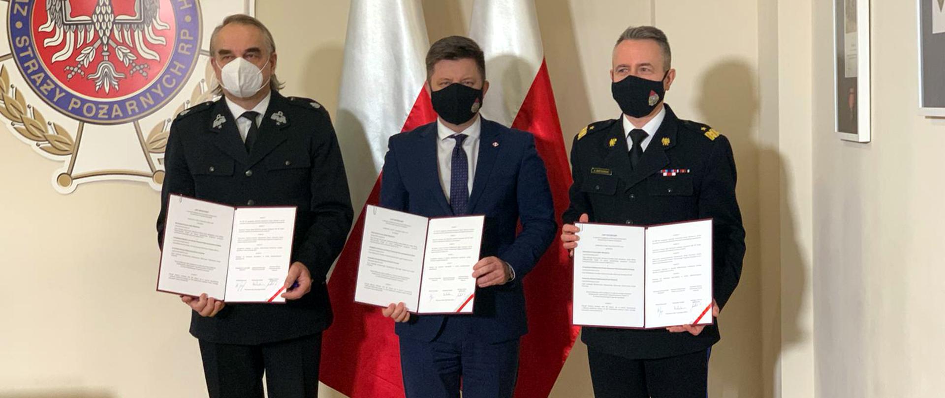 Zdjęcie przedstawia 3 stojących mężczyzn. Pierwszy z lewej w mundurze galowym Ochotniczej Straży Pożarnej, drugi w garniturze, trzeci w mundurze galowym Państwowej Straży Pożarnej. Wszyscy mężczyźni trzymają w dłoniach teczki z dokumentem w formacie A4. W tle znajdują się 2 flagi RP. 
