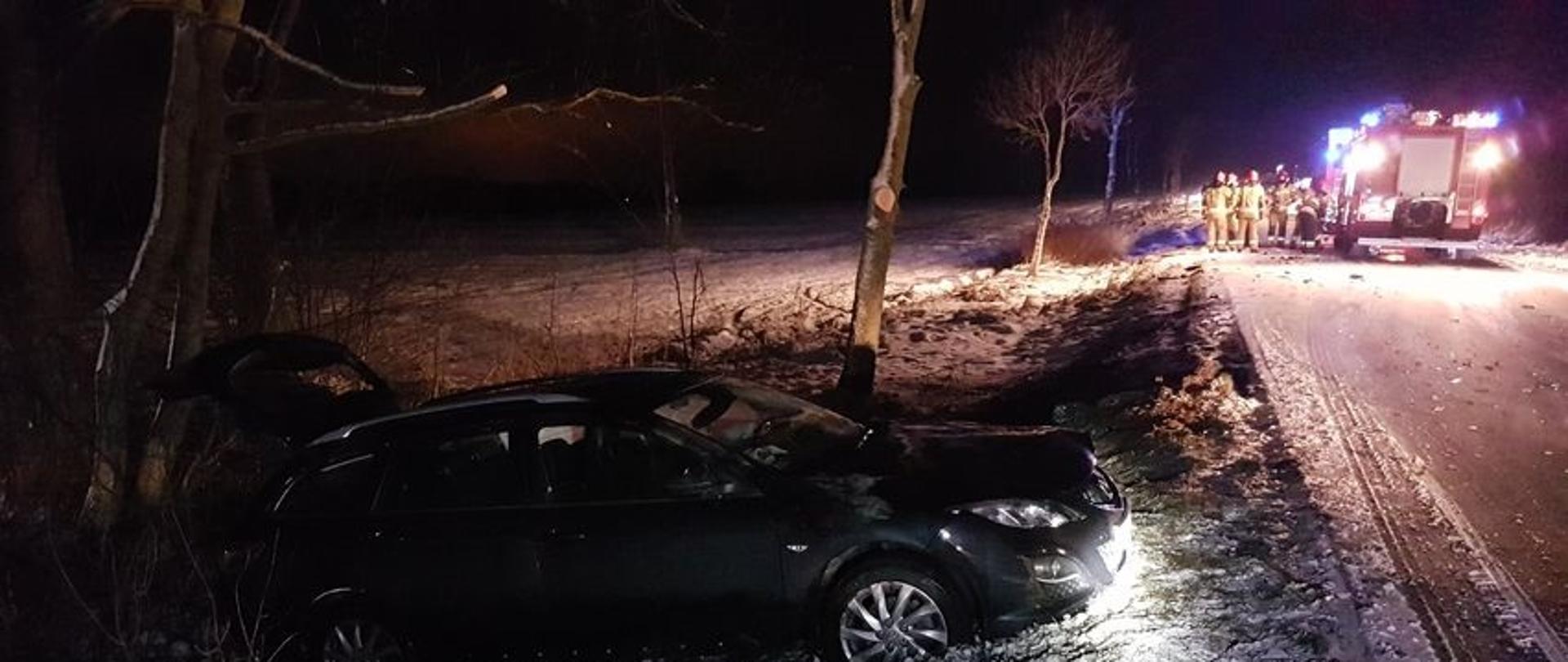 Zdjęcie przedstawia samochód osobowy marki Mazda koloru czarnego bokiem od strony pasażerów. Przód pojazdu skierowany w prawą stronę zdjęcia. Auto znajduje się na poboczu drogi z prawej strony widoczna droga, a na niej zastęp strażaków ubranych w ubrania specjalne obok samochodu strażackiego. Nocna, zimowa sceneria zdjęcia, widoczny śnieg zalegający na drodze i poboczu. Wrak samochodu na tle pojedynczych drzew