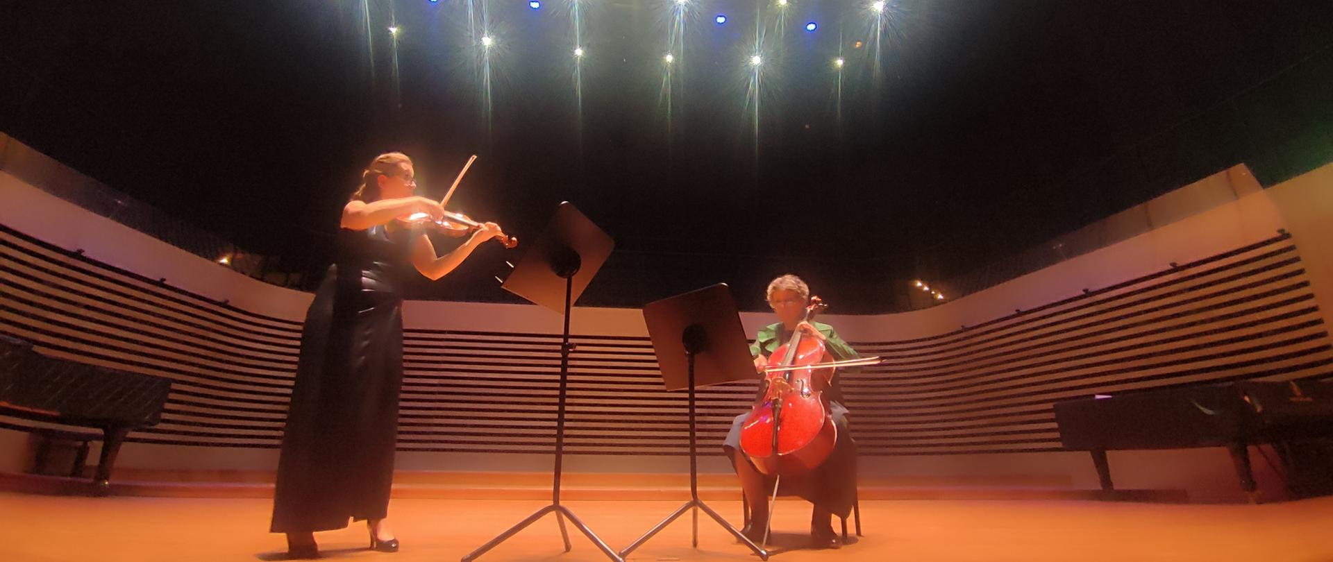 Po lewej stronie kobieta stoi przy pulpicie i gra na skrzypcach, obok po prawej stronie kobieta siedzi na krześle i gra na wiolonczeli, przed nią stoi pulpit.