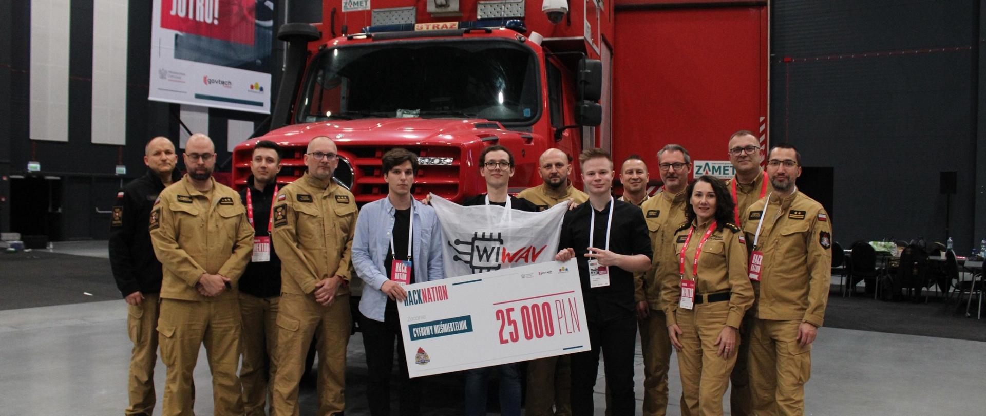 Grupa funkcjonariuszy PSP wraz z młodymi zwycięzcami hackathonu stoi przed pojazdem 220[B]90; zwycięzcy trzymają symboliczny czek na 25 000 zł i flagę zespołu projektowego.