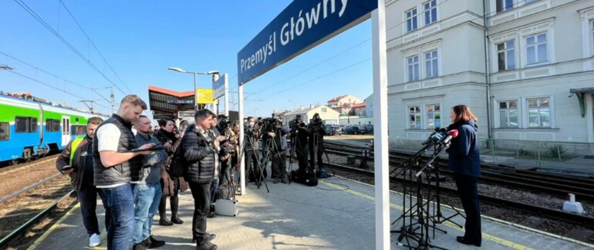 Wojewoda podkarpacki Ewa Leniart zabiera głos na dworcu kolejowym w Przemyślu podczas konferencji prasowej dotyczącej transportu w ramach ruchu migracyjnego. Przed wojewodą stoją dziennikarze, za nimi fragment pociągu