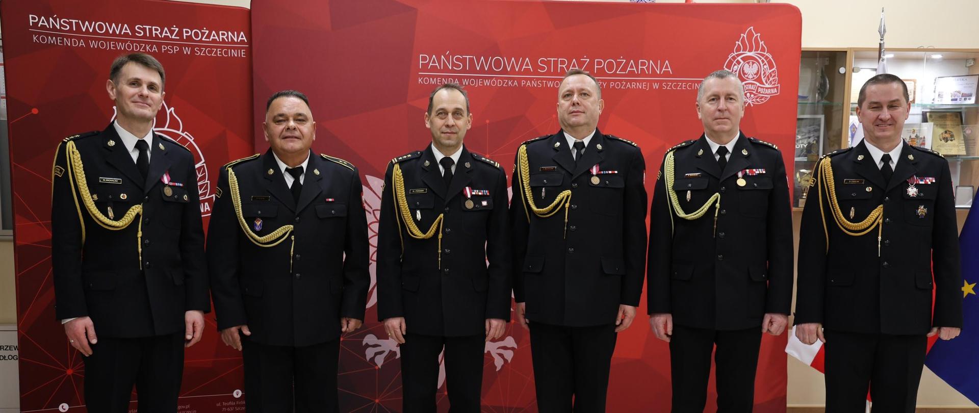 Sześciu strażaków w galowych, czarnych mundurach stoi w rzędzie na tle czerwonej ścianki z napisem Państwowa Straż Pożarna.