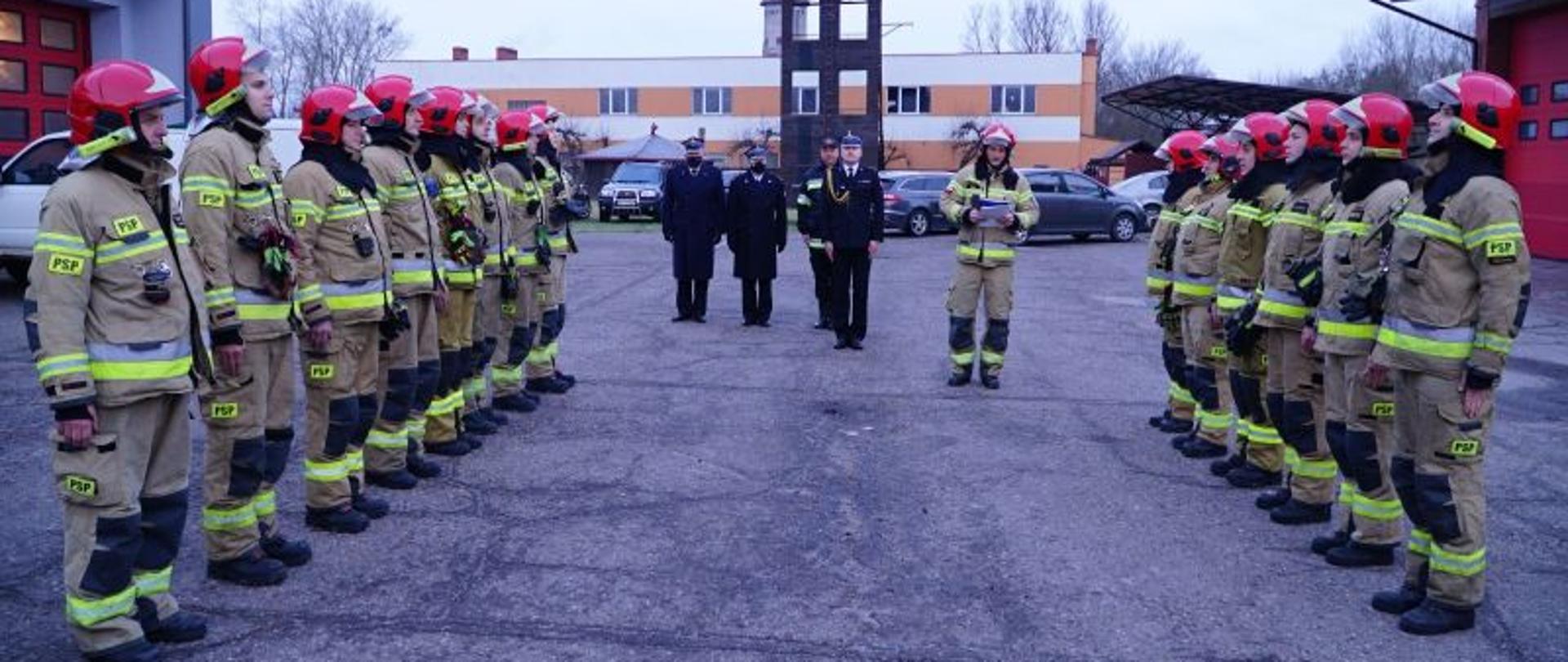 Zdjęcie przedstawia zmianę służby. Centralny punkt zdjęcia stanowią zmiany służbowe, które stoją na przeciwko siebie. Na drugim planie kadra dowódcza prowadząca zmianę służby oraz strażak odczytujący rozkaz dzienny