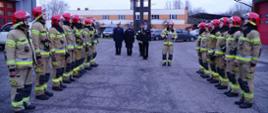 Zdjęcie przedstawia zmianę służby. Centralny punkt zdjęcia stanowią zmiany służbowe, które stoją na przeciwko siebie. Na drugim planie kadra dowódcza prowadząca zmianę służby oraz strażak odczytujący rozkaz dzienny