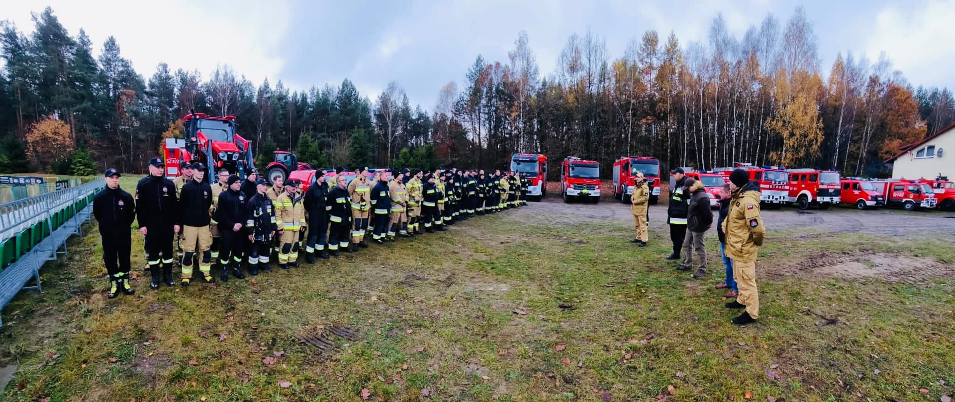 Zdjęcie pamiątkowe strażaków uczestniczących w ćwiczeniach, w tle las i ustawione w szeregu samochody pożarnicze.