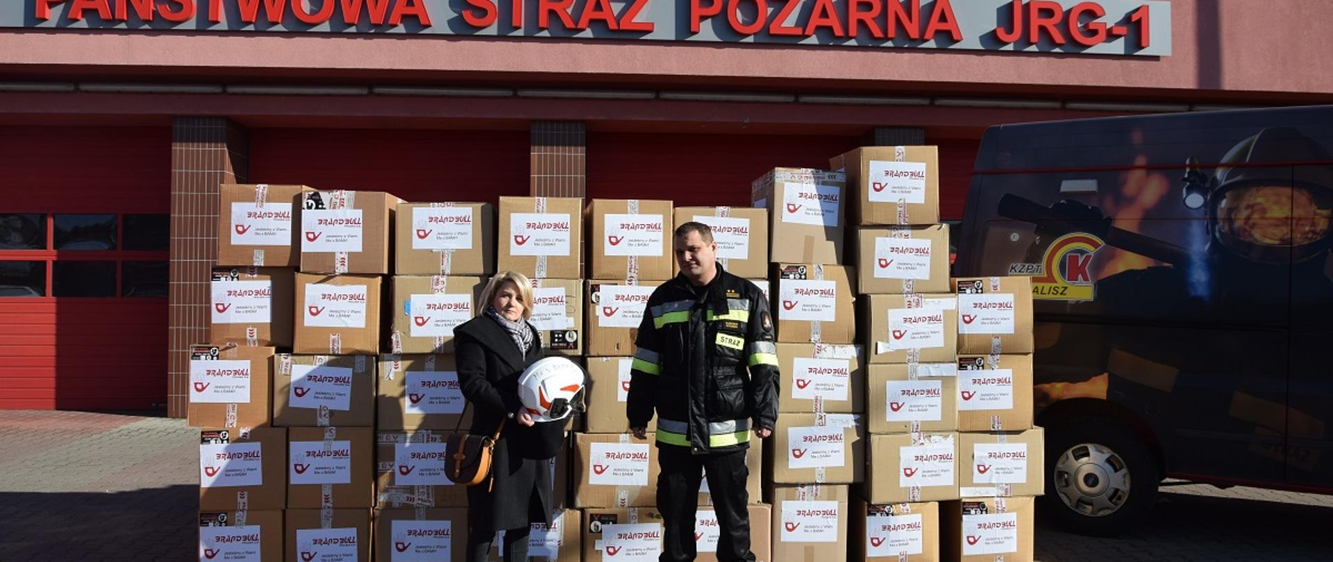 Na zdjęciu widać budynek JRG 1 z napisem czerwonym: Państwowa Straż Pożarna – JRG 1. Przed garażami kartony ze sprzętem. Przed kartonami Komendant Miejski PSP w Kaliszu oraz przedstawicielka firmy. Pora dnia dzień. Osoby stoją na kostce brukowej.