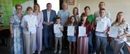 Sześcioro dzieci – Laureatów etapu regionalnego konkursu plastycznego dla dzieci „Wioski bez troski” trzymający pamiątkowe dyplomy, stojący w sali konferencyjnej Starostwa Powiatowego w Łosicach wraz z rodzicami i przedstawicielami organizatorów. W tle z lewej strony stoją baner prewencyjny KRUS i baner z logiem Starostwa Powiatowego w Łosicach, na ścianie z tyłu godło Polski.