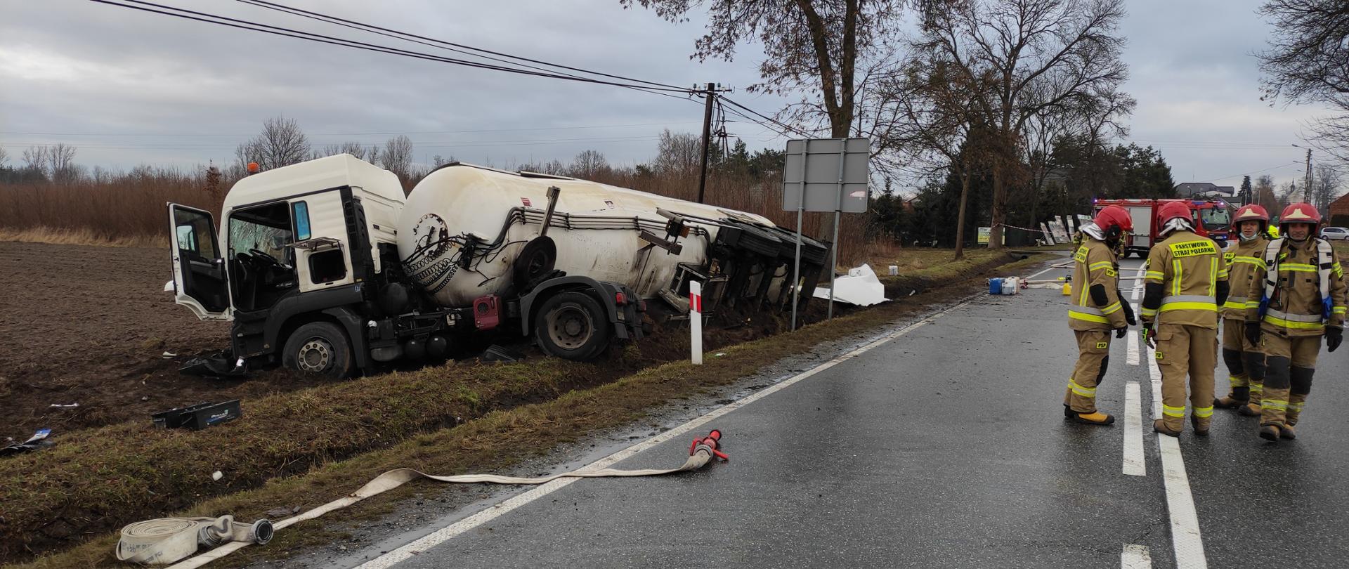 W rowie przewrócona biała cysterna wraz z ciągnikiem siodłowym, na drodze asfaltowej strażacy w ubraniach specjalnych z tył samochód pożarniczy.