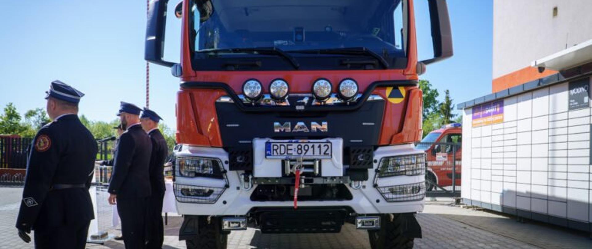 Zdjęcie przedstawia przód wozu strażackiego i trzech strażaków stojących bokiem