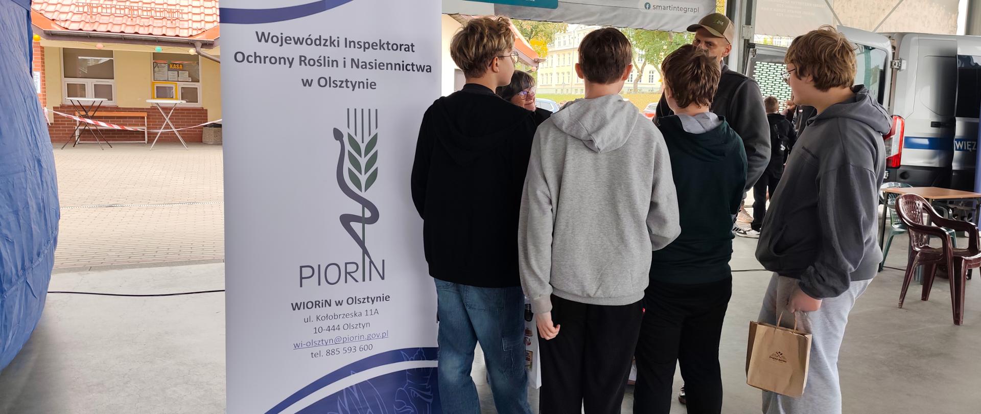 grupa młodzieży rozmawia przy stoisku Wojewódzkiego Inspektoratu Ochrony Roślin i Nasiennictwa w Olsztynie. Na pierwszym planie widoczny jest baner informacyjny PIORiN, a w tle znajdują się stoiska i reklamy wystawców