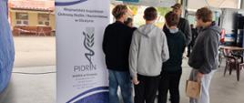 grupa młodzieży rozmawia przy stoisku Wojewódzkiego Inspektoratu Ochrony Roślin i Nasiennictwa w Olsztynie. Na pierwszym planie widoczny jest baner informacyjny PIORiN, a w tle znajdują się stoiska i reklamy wystawców