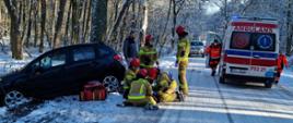widok na zimową ośnieżoną drogę, pięciu strażaków zajmuje się poszkodowaną osobą, która leży obok rozbitego auta, ich pracy przygląda się osoba postronna, z zaparkowanej obok karteki ratunkowej wybiegają ratownicy medyczni